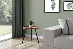 Jual Furnishings Siena lamp table displayed individually in a modern living room. The table features a round black marble top with white veining, a walnut-finished wooden rim, and slender black metal legs. It is styled with a small potted plant on top, next to a light grey corner sofa. The setting includes a sage green wall with framed botanical prints, floor-length green curtains, and a view of a garden through a large window.