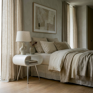 A styled bedroom scene featuring a bed dressed in neutral beige linens, a matching nightstand with a white ceramic lamp, and beige curtains framing a window in a softly lit room.