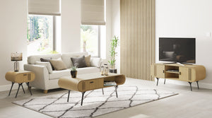 Modern living room featuring the Jual Furnishings Milano Collection, including an oak coffee table, side table, and TV stand with sleek black metal legs. The cohesive set complements a neutral sofa, decorative cushions, and a patterned rug, creating a warm and contemporary ambiance. Large windows allow natural light to enhance the minimalist aesthetic.