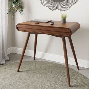 Jual Furnishings PC810 Walnut Desk, a modern curved-edge desk with a rich walnut finish, elegantly styled in a home setting. The desk features four tapered wooden legs and a discreet front drawer, complemented by a minimalist workspace setup with a laptop, plant, and accessories.