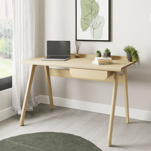Jual Furnishings PC811 Oak Desk styled in a modern home office setup. Features a sleek oak finish, a raised curved back panel, and a discreet drawer. Decorated with a laptop, books, plants, and minimalist artwork for a cozy workspace vibe.